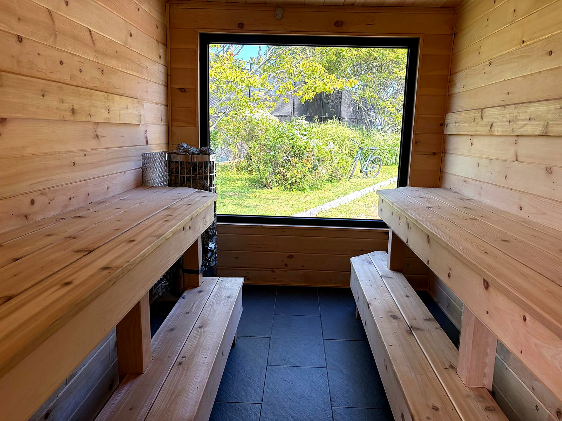 Cedar sauna interior with two-tier facing benches, stone heater and a large garden-view window